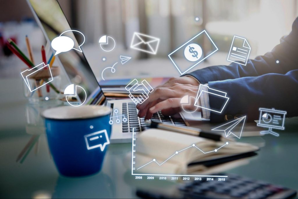 Manos sobre un portátil con iconos flotantes de marketing digital (email, gráficos, símbolo de dólar, chat, flechas de crecimiento) y taza de café azul. Ubicada bajo el H2 "Cuánto invertir según tu sector"