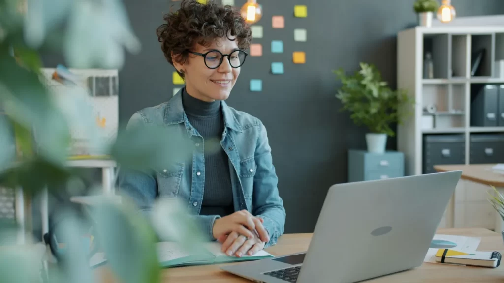 Mujer sonriendo ante su portátil en una oficina luminosa con notas adhesivas en la pared