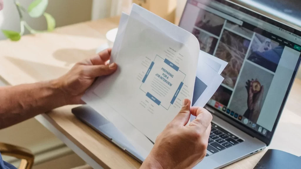 Persona revisando documentos impresos junto a un portátil en un espacio de trabajo luminoso